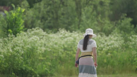 A-rear-view-of-a-middle-aged-casually-looking-woman,-wearing-a-knitted-dress-and-a-sun-hat,-on-a-stroll-in-a-park-Her-dark-hair-gathered-in-a-ponytail