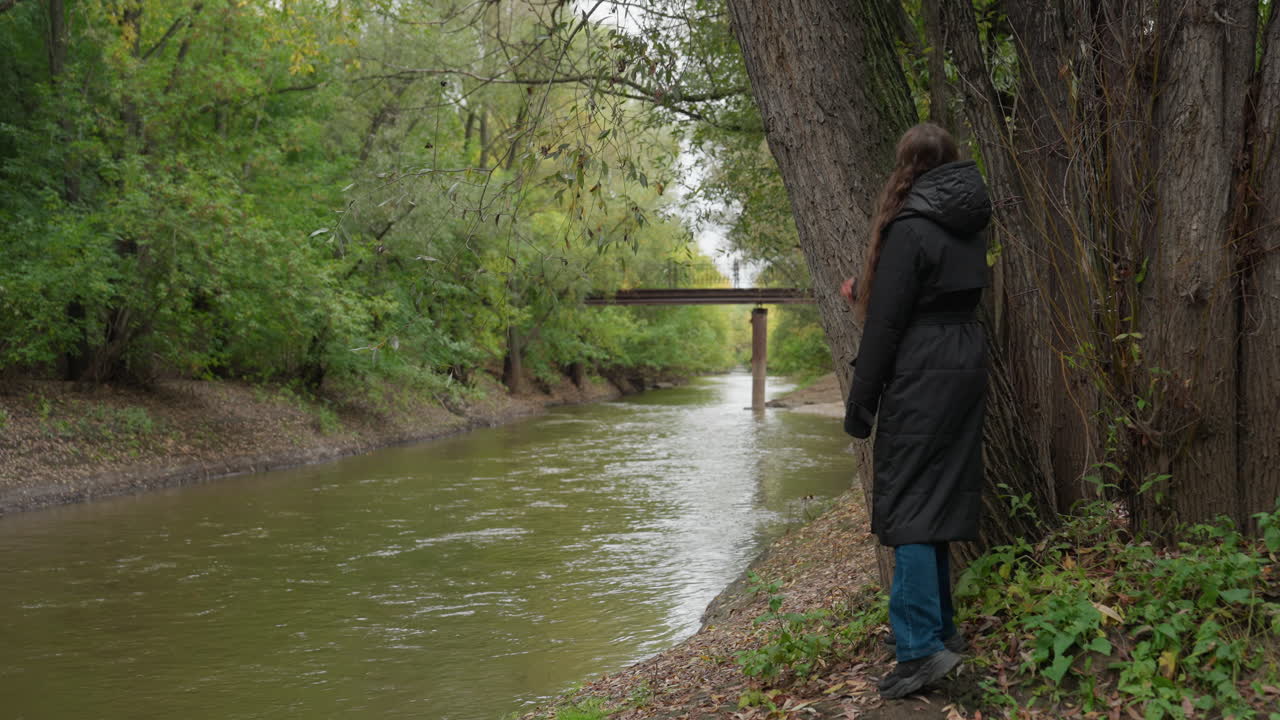 Persona che cammina lungo la riva del fiume sotto un cielo nuvoloso, con cappotto scuro e jeans, che si ferma a toccare il tronco muschioso di un albero accanto a un fiume lento, ponte in lontananza incorniciato dal fogliame autunnale, atmosfera riflessiva e calma