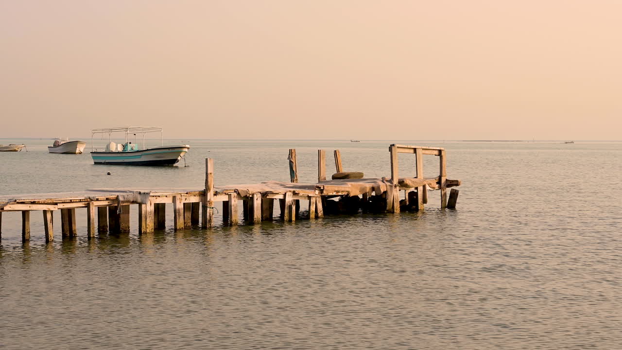 Wooden old pier on beautiful peaceful calm sea water waves with boats in background over sky, Bahrain