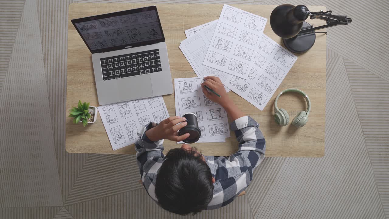 vista superior de un artista masculino asiático bebiendo café mientras dibuja un guión gráfico para la película en la mesa con una computadora portátil y auriculares en el estudio