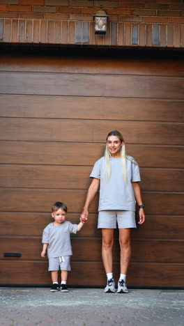 long-haired caucasian woman takes a hand of a toddler boy standing nearby. portrait of mom and son standing at the wooden wall outdoors. vertical video