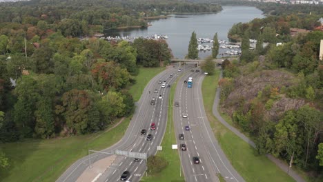 drone footage of cars and buses driving on road in stockholm, sweden on overcast day with green grass and trees and with a lake in the background on the start of fall