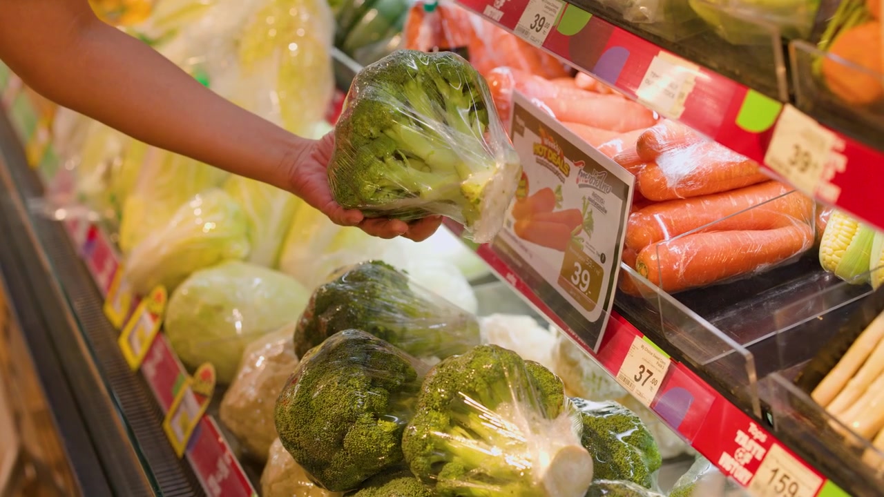 Primo piano di una mano che prende broccoli confezionati in plastica da uno scaffale di un supermercato a Bangkok.