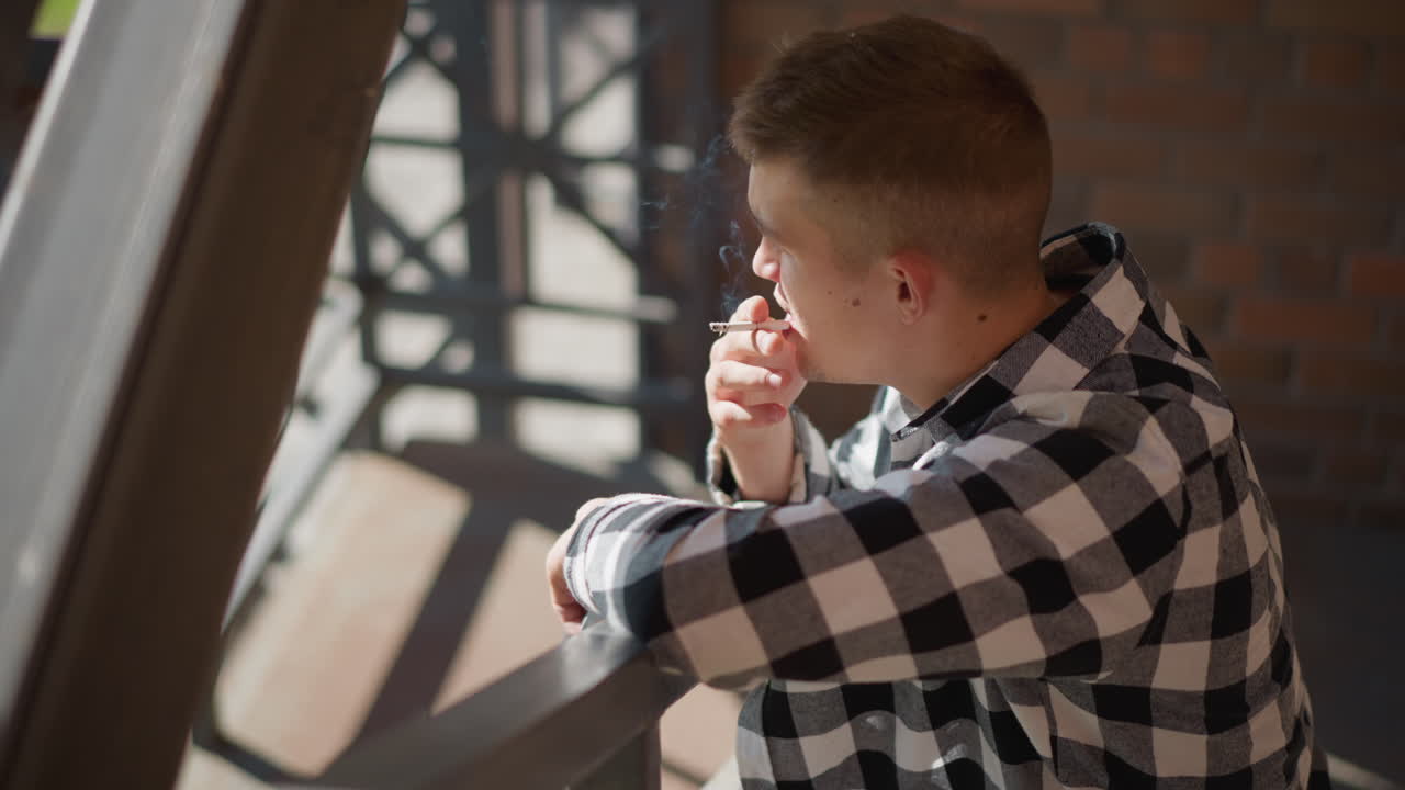 aerial angle of young man wearing checkered shirt leaning on railing and smoking while exhaling smoke that gently swirls upward in sunlight near brick wall and metal stair railings outdoors