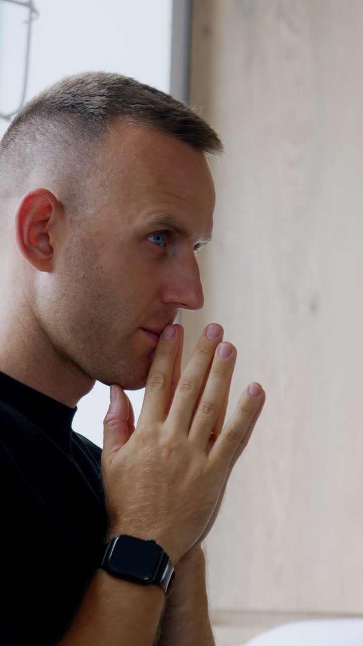 Tensed Caucasian man in black t-shirt standing at the window. Nervous man holding hands in front of his face. Husband worries for wife at childbirth. Vertical video
