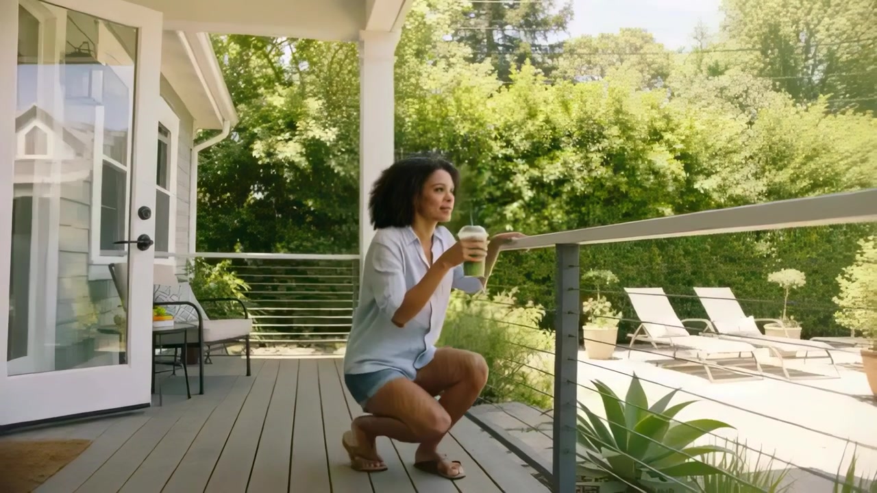 Stepping woman in shirt shorts exiting French doors to deck crouching rail sipping cup viewing pool