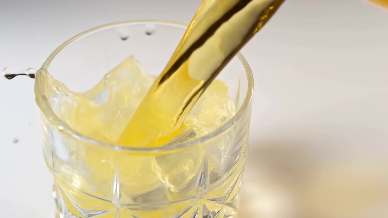 Tampilan dekat minuman keemasan yang dituangkan ke dalam gelas bening berisi es batu dengan latar belakang putih cerah, dilengkapi pencahayaan studio yang lembut dan konsep penyegar yang bersih