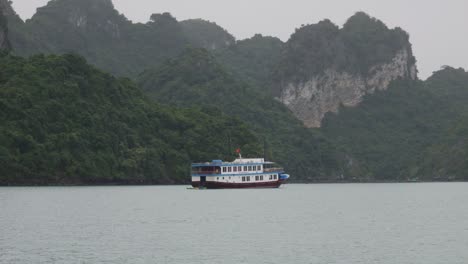 Cruise-ship-sailing-through-Ha-Long-Bay-in-Vietnam,-surrounded-by-mountains-and-captured-on-a-cloudy-day
