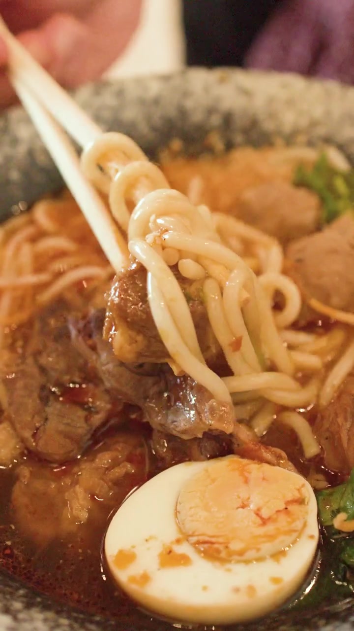 Primo piano di bacchette che sollevano spessi noodles di riso da una ciotola di zuppa di manzo piccante a Bangkok