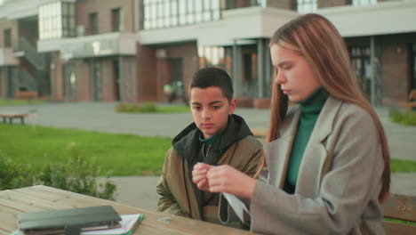 Little boy nods head attentively while watching aunt fold paper into plane at wooden table outdoors, both seated together with notebooks nearby in calm urban environment