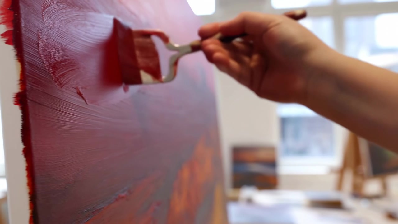 Rechterhand en onderarm reiken over gestructureerd gespannen canvas, brengen glanzende rode en oranje verf aan met kwast of paletmes, verspreiden pigment in levendige streken onder zacht natuurlijk tegenlicht
