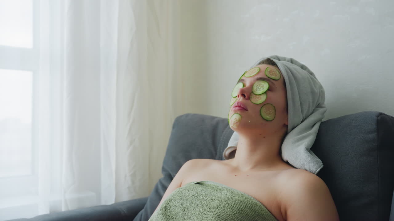 Woman relaxing and feeling comfortable, wrapped in towel with cucumber slices placed on her face, she enjoys a peaceful, rejuvenating skincare routine with eyes closed, embracing the soothing effects