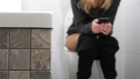a woman with trousers down sitting on the toilet and using a smartphone.