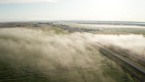 Aerial-view-of-fog-hanging-low-over-green-farmland-cut-by-highway-with-traffic-moving-both-directions