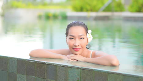 close-up of an exotic woman with wet hair and a flower behind her left ear leans along the edge of a resort swimming pool