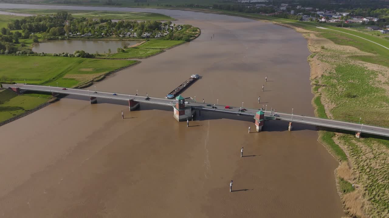 Aerial orbit showing the Jann-Berghaus Bridge and a cargo vessel navigating the Ems River in Leer, Germany. Road traffic, rural landscape, and calm river conditions captured in evening light.