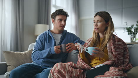 Sick lady feeling headache talking husband at sofa. Smiling man drinking hot tea