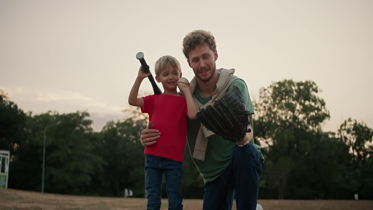 Portrait of a Happy blond boy in a red T-shirt who holds a baseball bat and stands with his dad in a Green T-shirt with curly hair and stubble and posing in the park