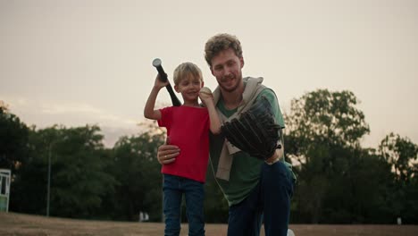 Portrait-of-a-Happy-blond-boy-in-a-red-T-shirt-who-holds-a-baseball-bat-and-stands-with-his-dad-in-a-Green-T-shirt-with-curly-hair-and-stubble-and-posing-in-the-park