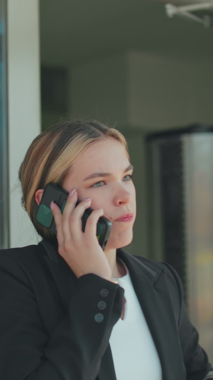 Frustrated woman during phone call lowers phone briefly, continues conversation after work while exiting office building, dressed in formal attire, glass door reflects cityscape