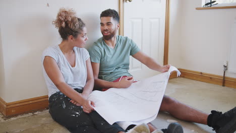 couple sitting on floor looking at floor plans in empty room of new home