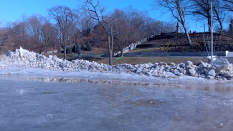 Drohnenschwenkaufnahme-Eines-Parks-Am-Rande-Eines-Zugefrorenen-Flusses-Mit-Haufen-Von-Eisfragmenten-Und-Trümmern-Am-Flussufer