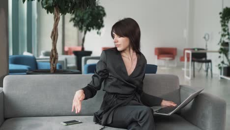 young, beautiful woman in a suit plays music on her laptop and begins to dance joyfully, raising her hands up while sitting on the couch