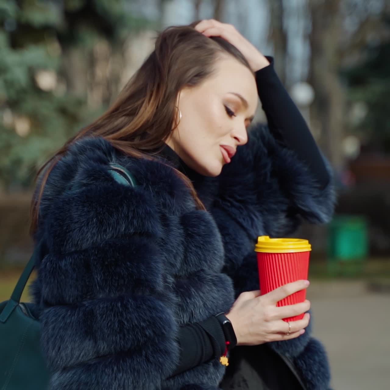 Side view of a beautiful woman outdoors. Pretty girl in luxury fur coat walking in the park with a plastic cup of coffee. Slow motion.