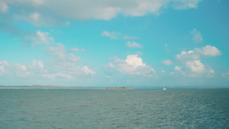 a small island, sits in the center of a tropical blue ocean on a beautiful sunny day, whilst fluffy white clouds float overhead and a small boat sails poa