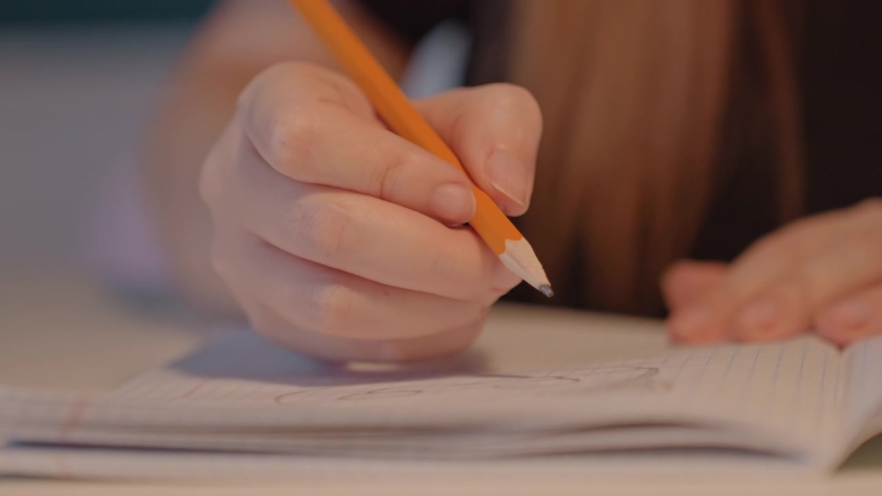 Primo piano di una giovane donna che scrive con una matita arancione. La sua mano sinistra poggia vicino a un libro mentre la donna disegna creando linee. La scena è ambientata al chiuso, a una scrivania o a un tavolo, con un'illuminazione soffusa che enfatizza i dettagli