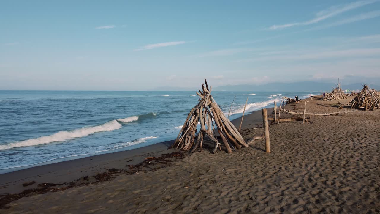 Langsom filmisk luftdroneoptagelse af en træ-drivtømmer tipi, der flyver væk fra en sandstrand ved kysten nær Alberese i den ikoniske Maremma naturpark i Toscana, Italien, med blå bølger