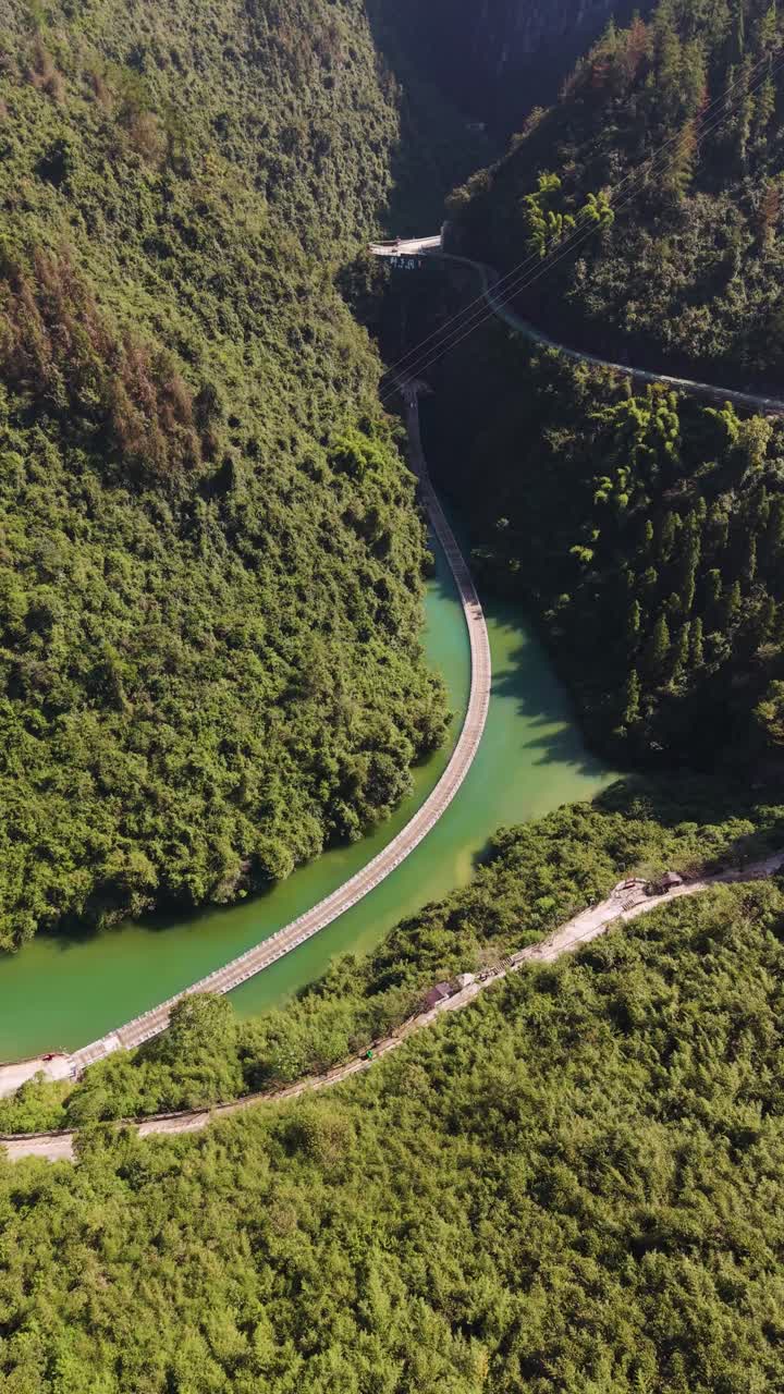 Vertical aerial view of the Shiziguan Floating Bridge over a turquoise river in Hubei Province, China. The narrow wooden bridge winds through a deep, lush green river gorge