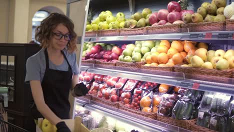 Close-up-of-pretty-caucasian-worker-in-black-apron-and-gloves-stocking-the-fruits-in-supermarket.-Young-employee-in-glasses-at