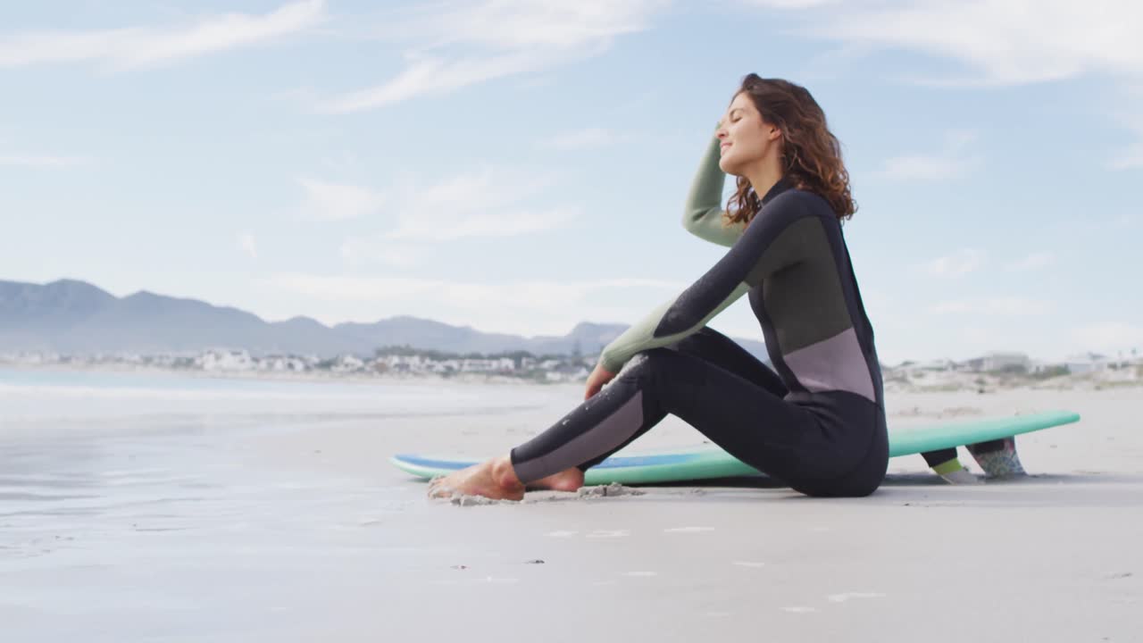 해변에 앉아 서프보드와 함께 바다를 바라보고 미소 짓는 행복한 혼합 인종 여성