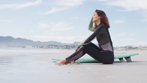 happy mixed race woman sitting on beach with surfboard looking out to sea and smiling