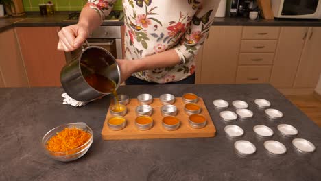 Slow-zoom-in-close-up-shot-of-a-young-woman-pouring-hot-a-yellow-orange-liquid-beeswax-balm-into-empty-circular-aluminum-containers