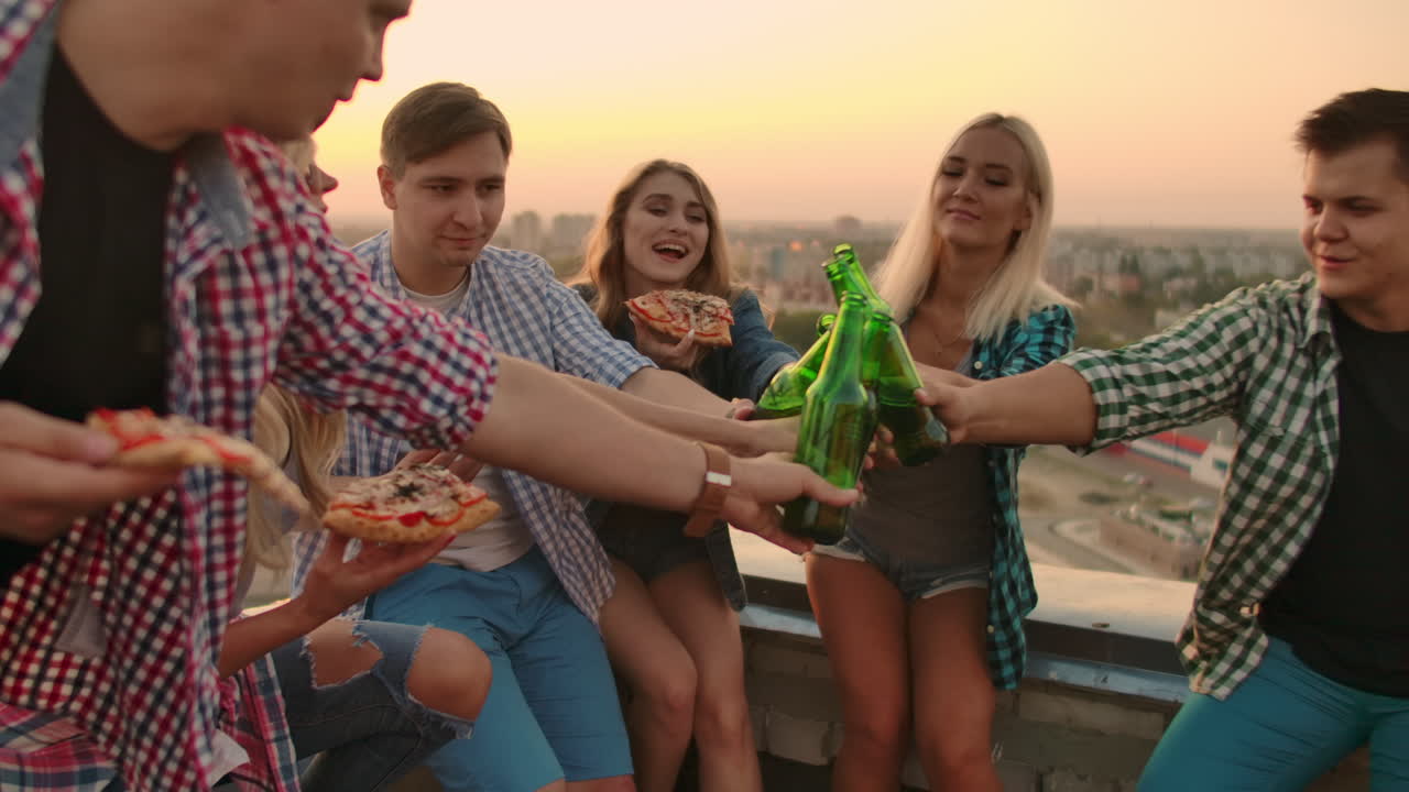 Young girls and boys clinks glasses and drinks beer from green bottels on the party with friends on the roof at the sunset.They are sitting together eat hot pizza after in summer everning.
