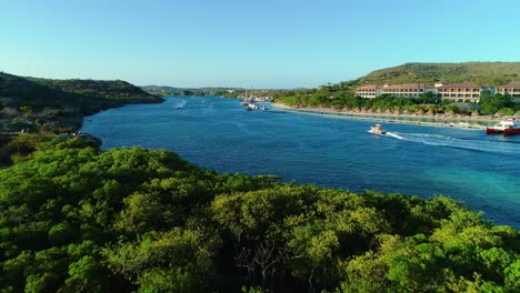 Boats-and-Jet-Ski-waverunners-passing-by-pier-and-beach-of-sandals-resort-curacao,-drone-dolly-above-channel