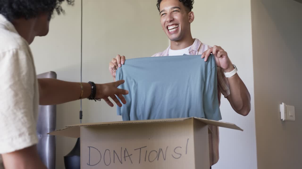 African American friends sorting clothes into box marked DONATIONS! presenting blue T-shirt at home