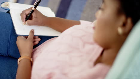 An-African-American-woman-is-writing-in-a-notebook-at-home,-wearing-a-pink-top-and-blue-jeans