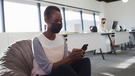 African-american-businesswoman-wearing-face-mask-and-using-smartphone-in-office