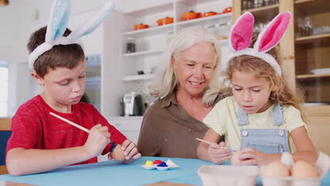 Großmutter-Mit-Enkelkindern,-Die-Hasenohren-Tragen-Und-Zu-Hause-Gemeinsam-Ostereier-Dekorieren