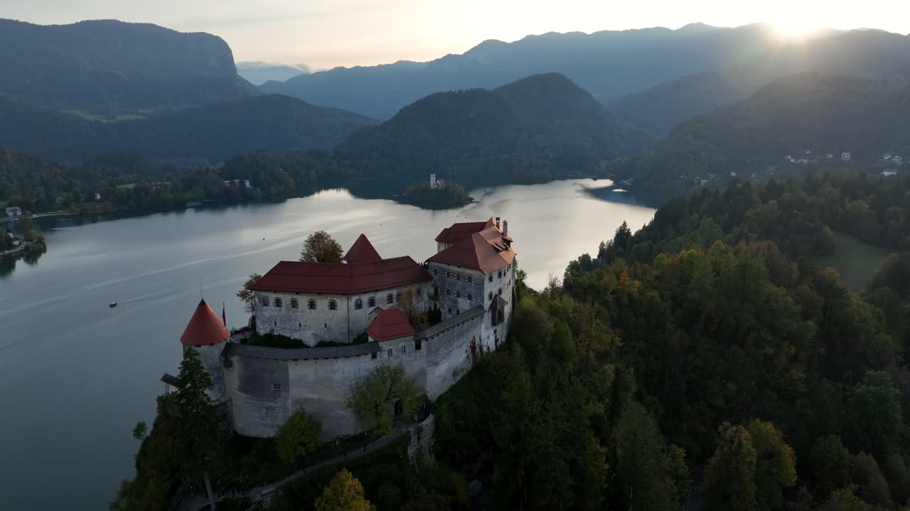 Ripresa cinematografica con drone dello storico Castello di Bled, appollaiato su una scogliera sopra il Lago di Bled, Slovenia. L'iconica chiesa sull'isola, le Alpi Giulie e l'illuminazione dell'ora d'oro sull'acqua.