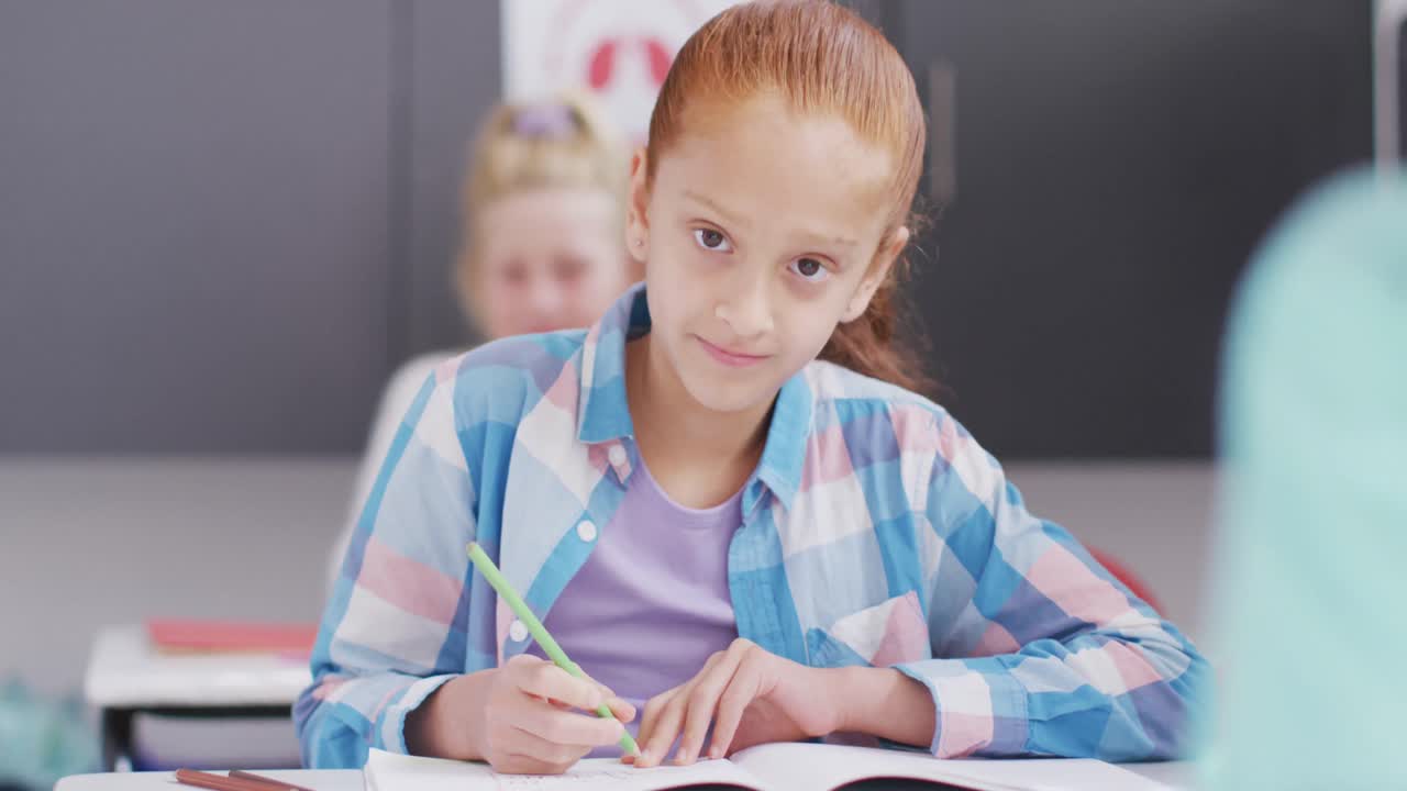retrato de una colegiala caucásica sentada en un escritorio sonriendo y escribiendo en el aula