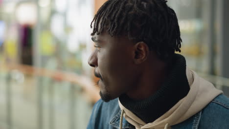 confident young man in denim jacket and hoodie sipping coffee from paper cup while looking ahead with calm expression, enjoying quiet moment in modern indoor space with soft background