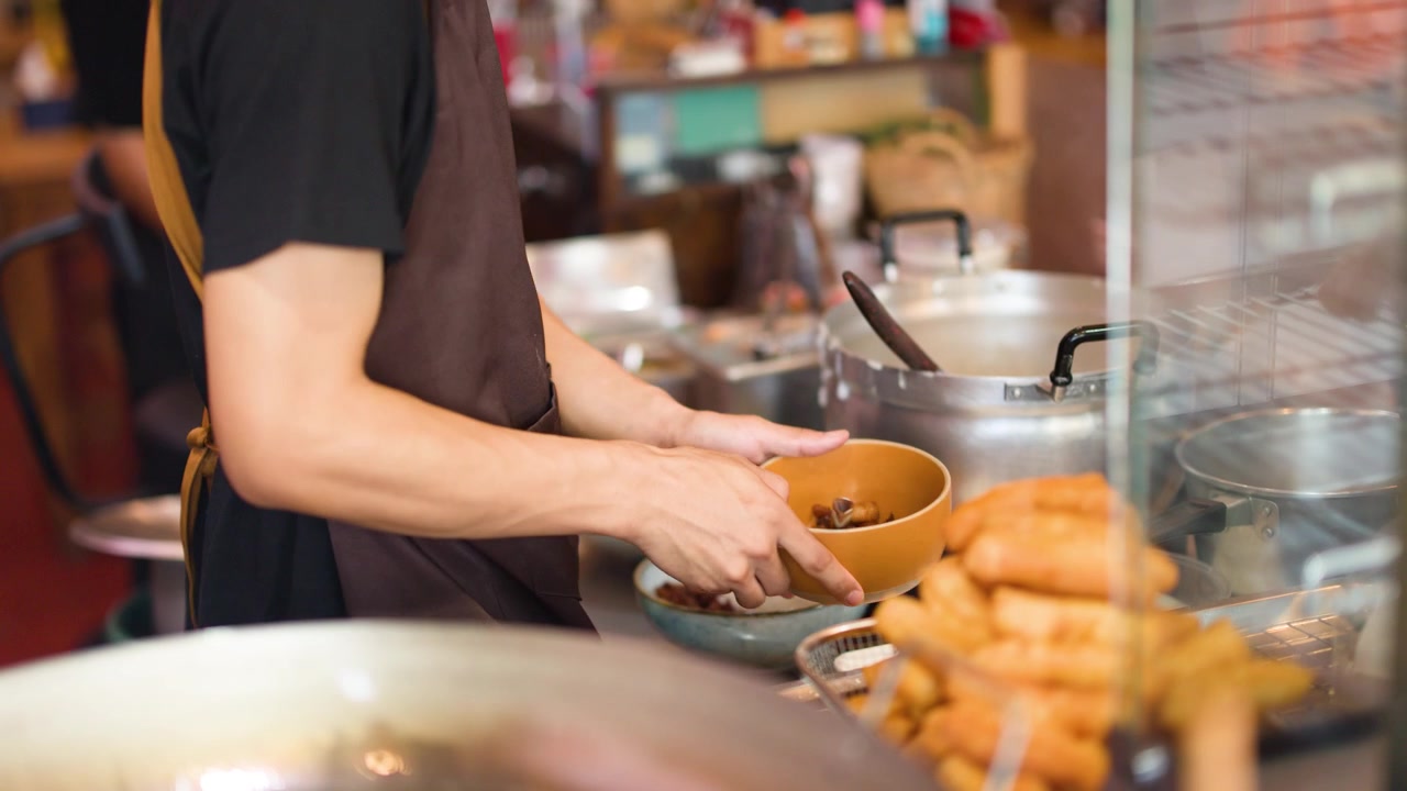 Asian male vendor ladles hot broth into bowls at a busy outdoor food stall.