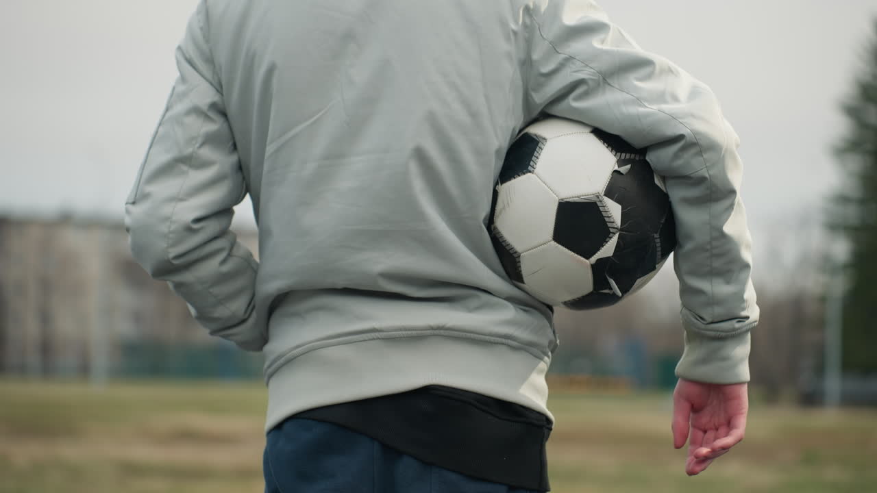 Back view of a person standing with a soccer ball tucked under the right arm, wearing a gray jacket with their left hand in the pocket, the background shows a blurred view of a grassy field