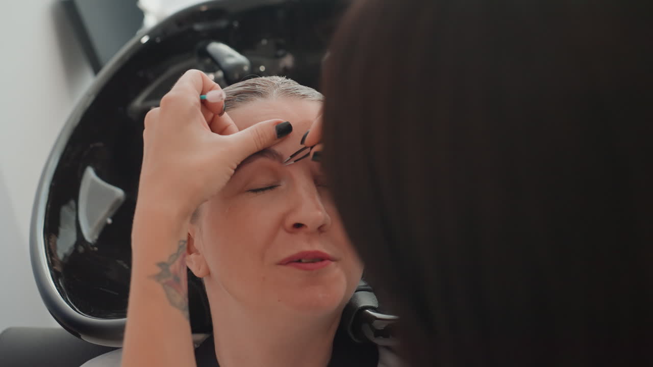 woman receiving eyebrow threading in salon, esthetician with tattooed hands shapes brows over basin, client reclining with closed eyes, precise hand movements, serene expression, warm lighting