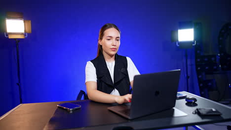 Brunette Caucasian lady wearing white t-shirt and vest sits at desk working at laptop. Soffits switched on are at the backdrop.