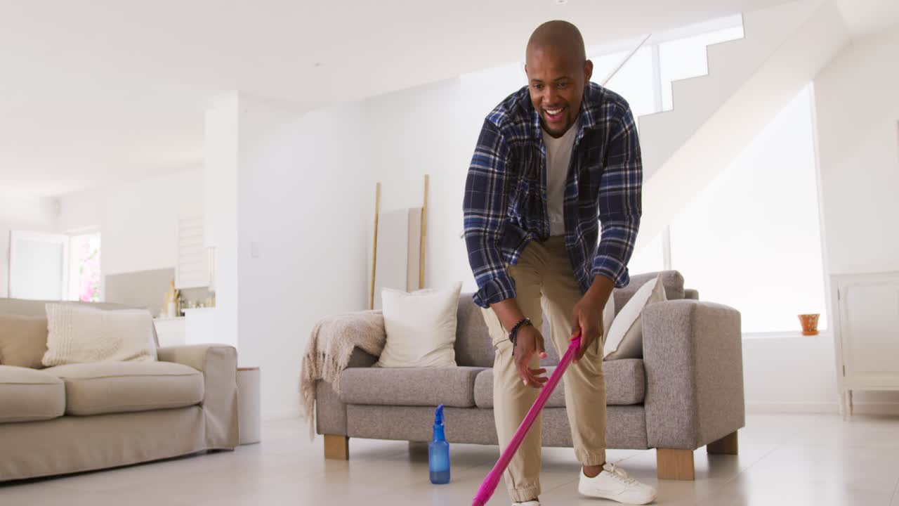 African American man in plaid spraying with blue bottle, pushing pink mop in living room smiling